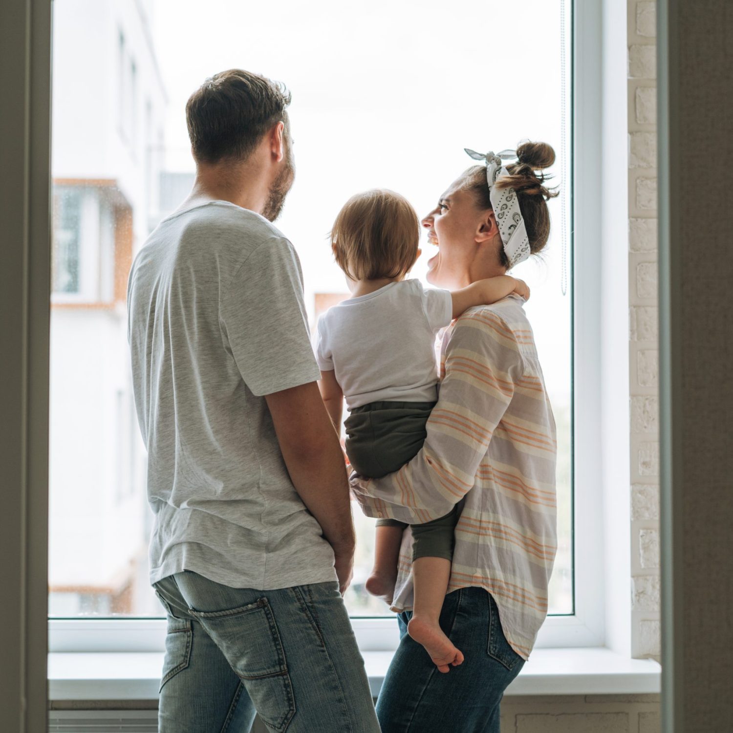 Young family man father and mother woman with baby girl on window sill looking at window at home